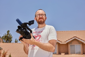 In a very bright, sunny driveway, a white man holds a video camera while squinting into the sun.