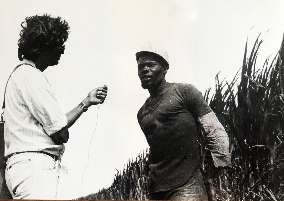 black and white photo from the Victor Jara Collective featuring a man holding a small microphone up to another man wearing a hard hat standing against a row of brush