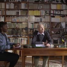 Alice Diop is a Senegalese-French woman wearing a multicolored jacket. She is sitting in a library, speaking to Pierre Bergounioux, an older white French male writer. They're both being filmed. From Alice Diop's 'Nous.' Photo by Sarah Blum. Courtesy of Mubi.