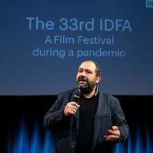 IDFA Artistic Director and filmmaker Orwa Nyrabia is a Syrian man with a beard and short hair. He is wearing a black jacket and a sweater while addressing the in-person and virtual audience at IDFA 2020. There is a screen behind him. Photo: Coen Dijkstra. Courtesy of IDFA