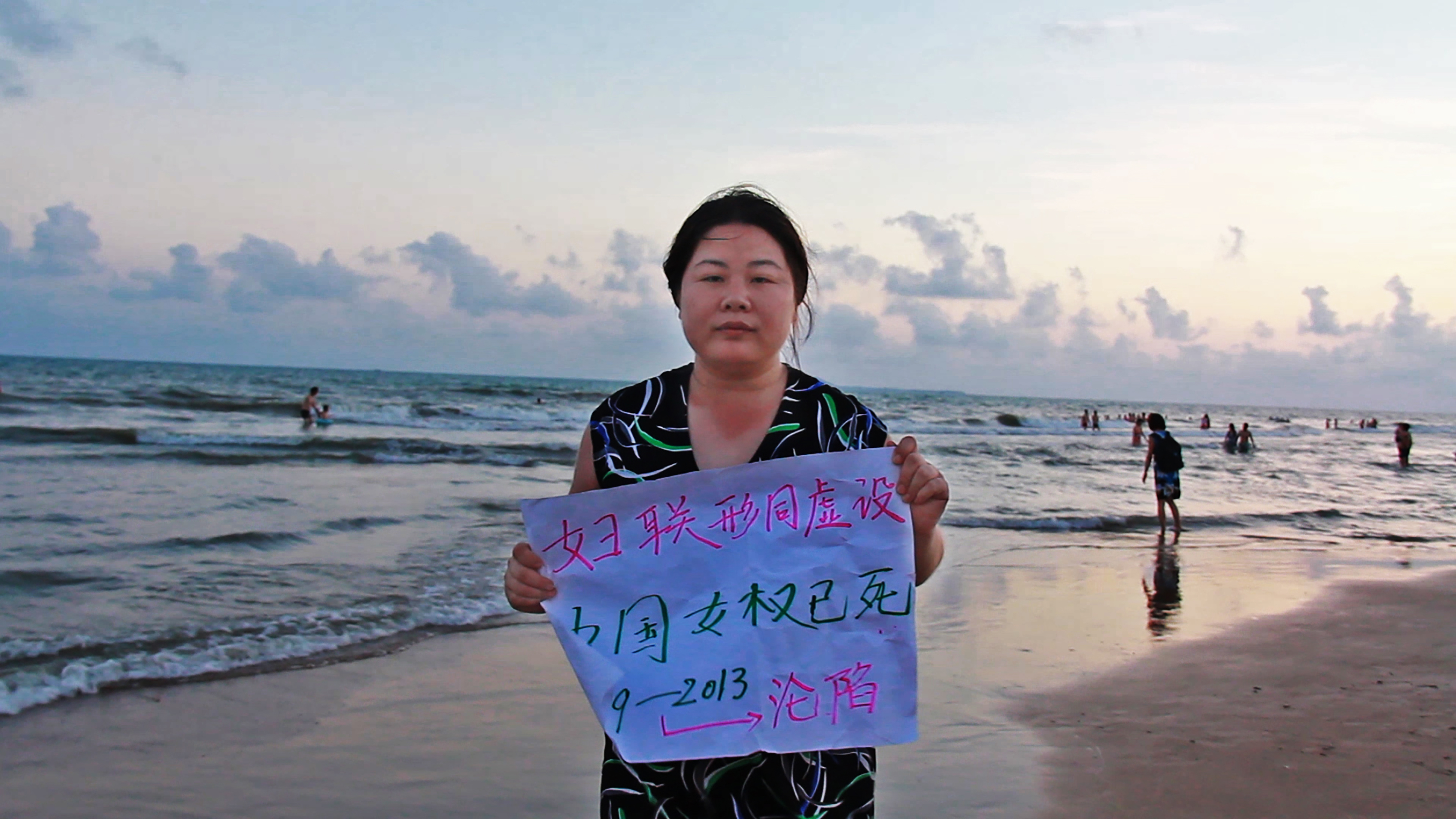 Still image from Nanfu Wang’s "Hooligan Sparrow." Ye Haiyan, a fair complexioned adult with black hair, stands at the beach wearing a black dress with blue, green, and white designs. She is holding a sign with simplified Chinese characters.