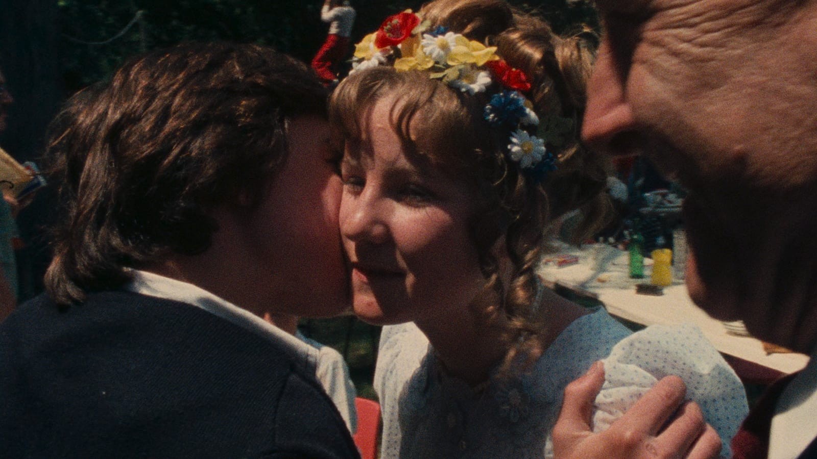Man kissing a woman on the cheek. Woman is wearing a white dress with red, yellow, white, and blue flower headband.