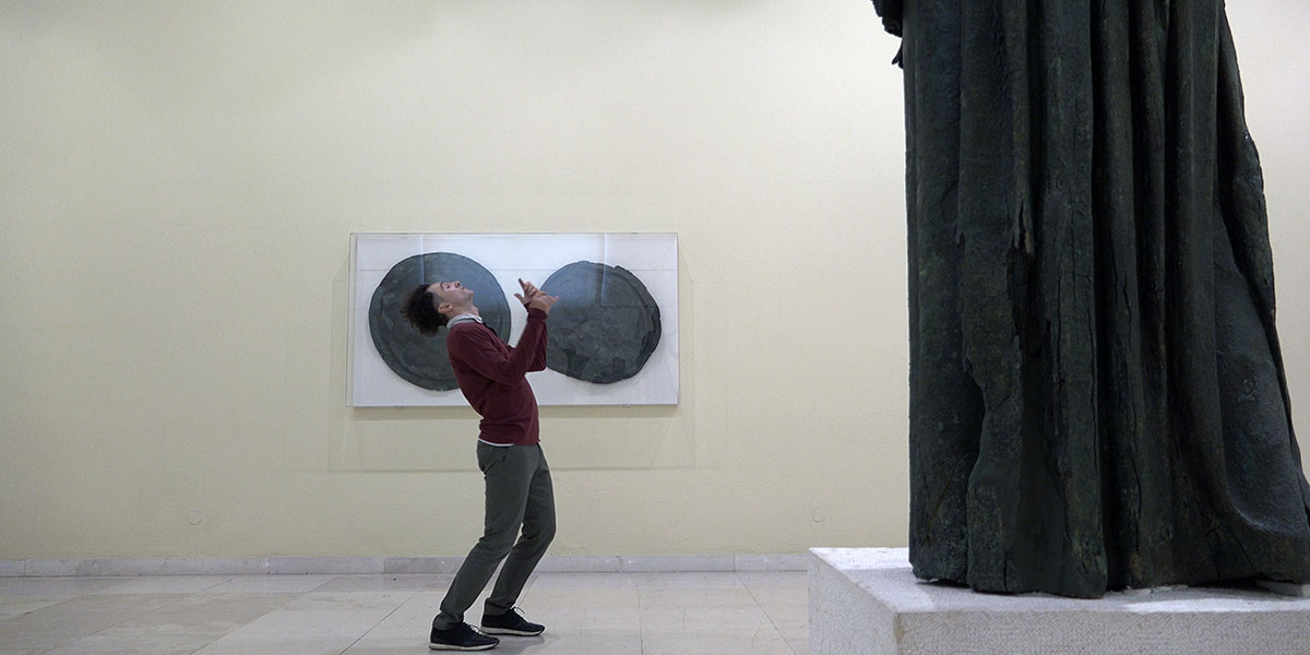 In front of a black monolithic statue, a man in a red sweater stands bent over backward.