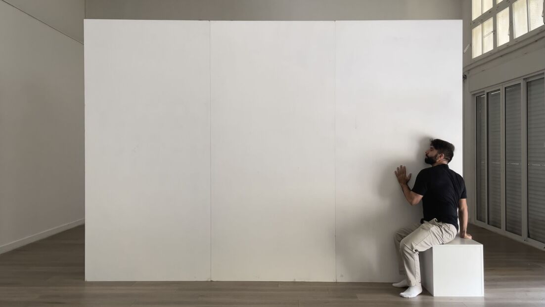 Alaa Minawi sits on a white box, with his ear pressed against a white wall in the center of a bare room.