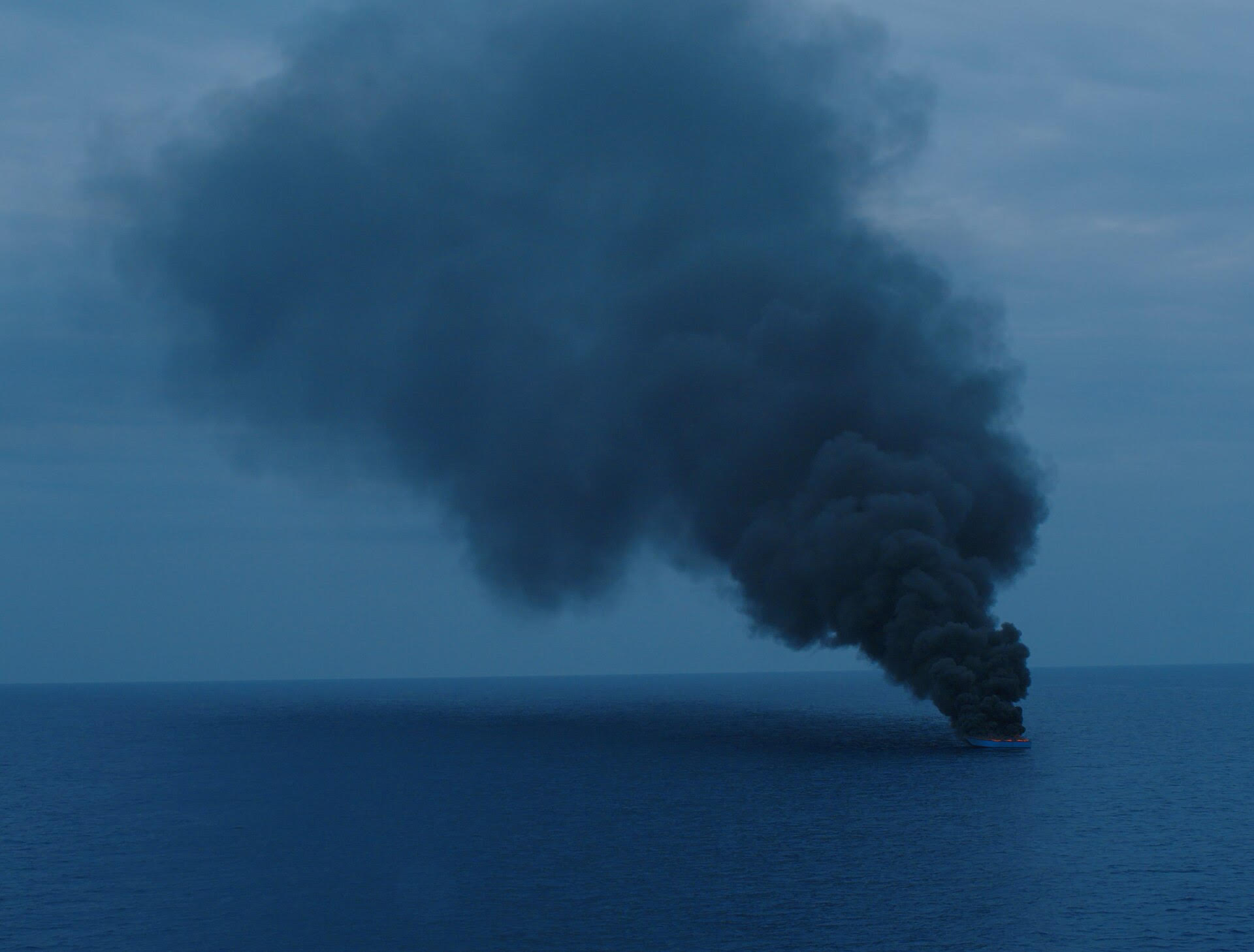 A boat in vast, open water billows black smoke into the night sky.