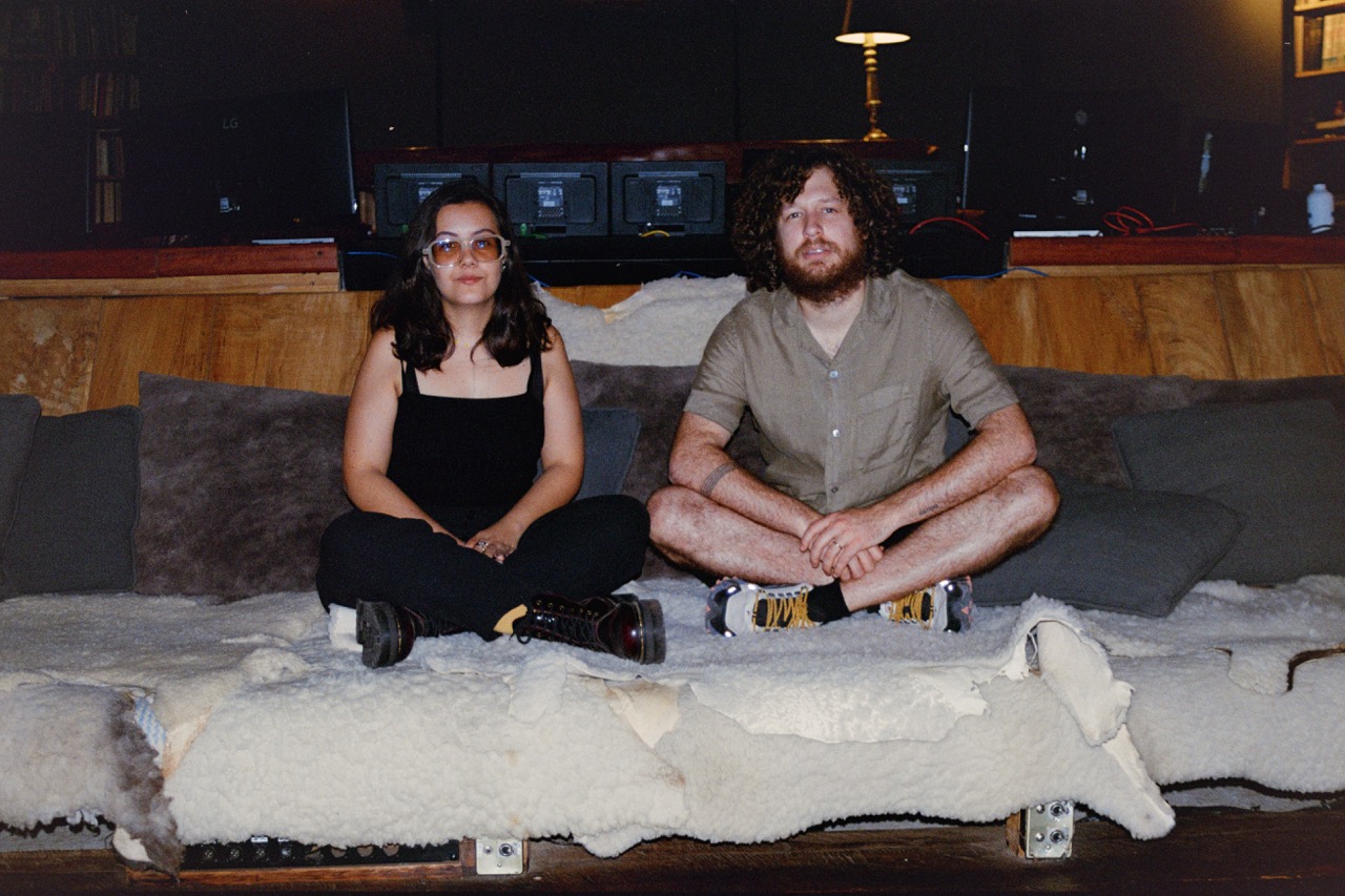 A brunette woman dressed in black sits next to a man with a beard on a couch covered in various furs. 