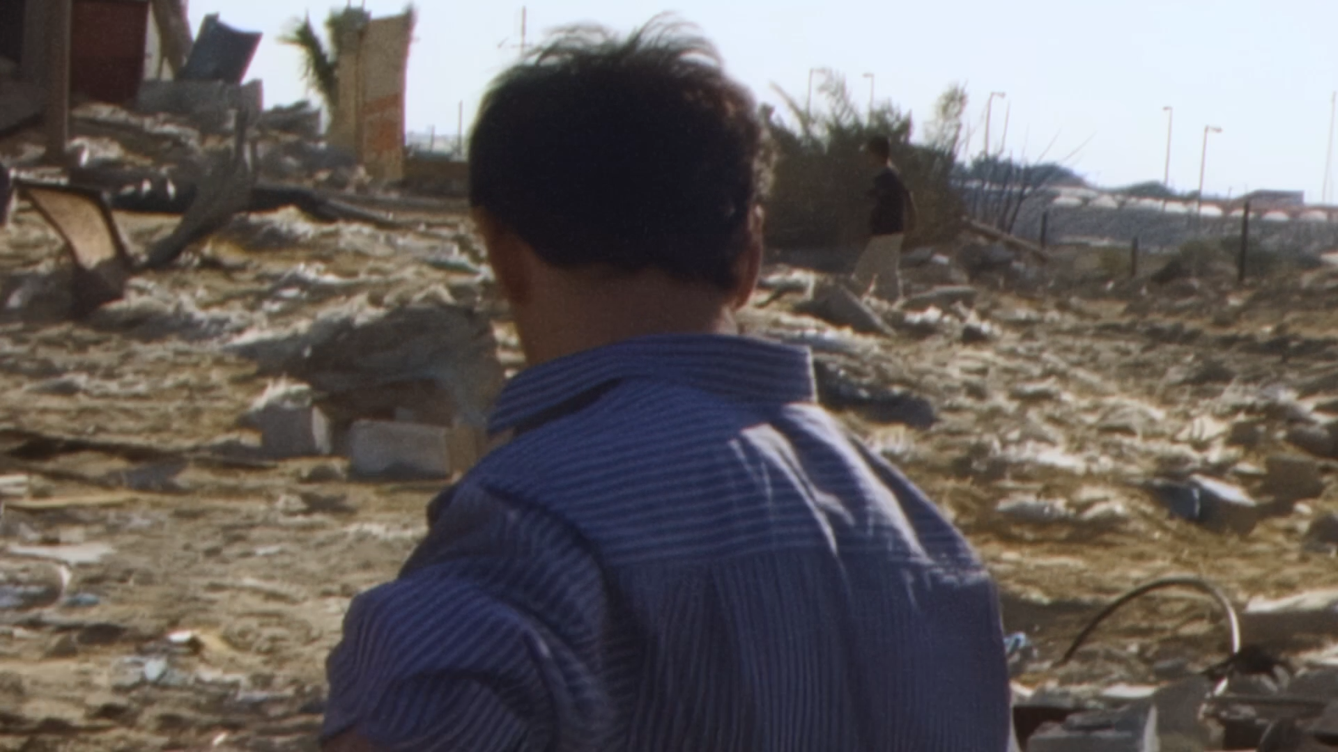 DV tape image of the back of a man's head, walking through a field with building rubble.
