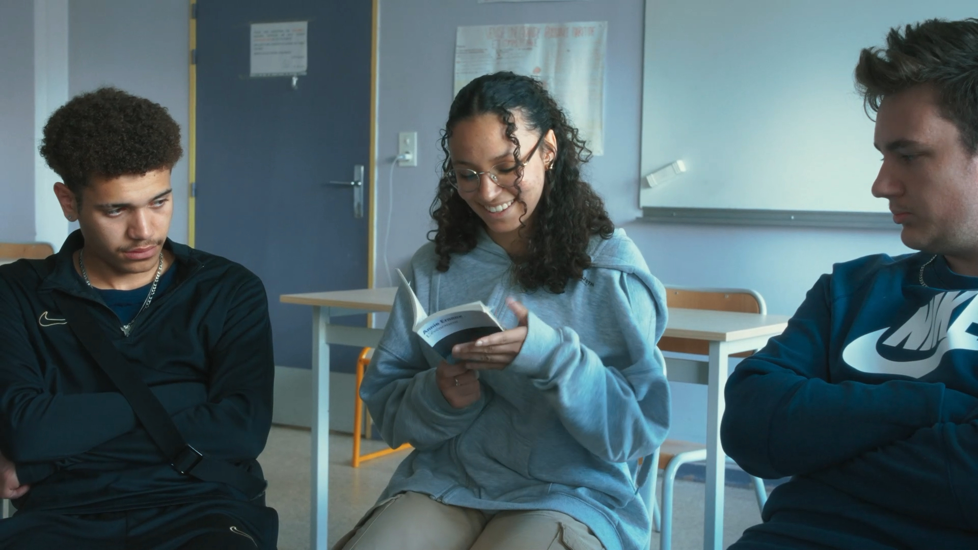 Three young people look at a book, while sitting in a classroom.