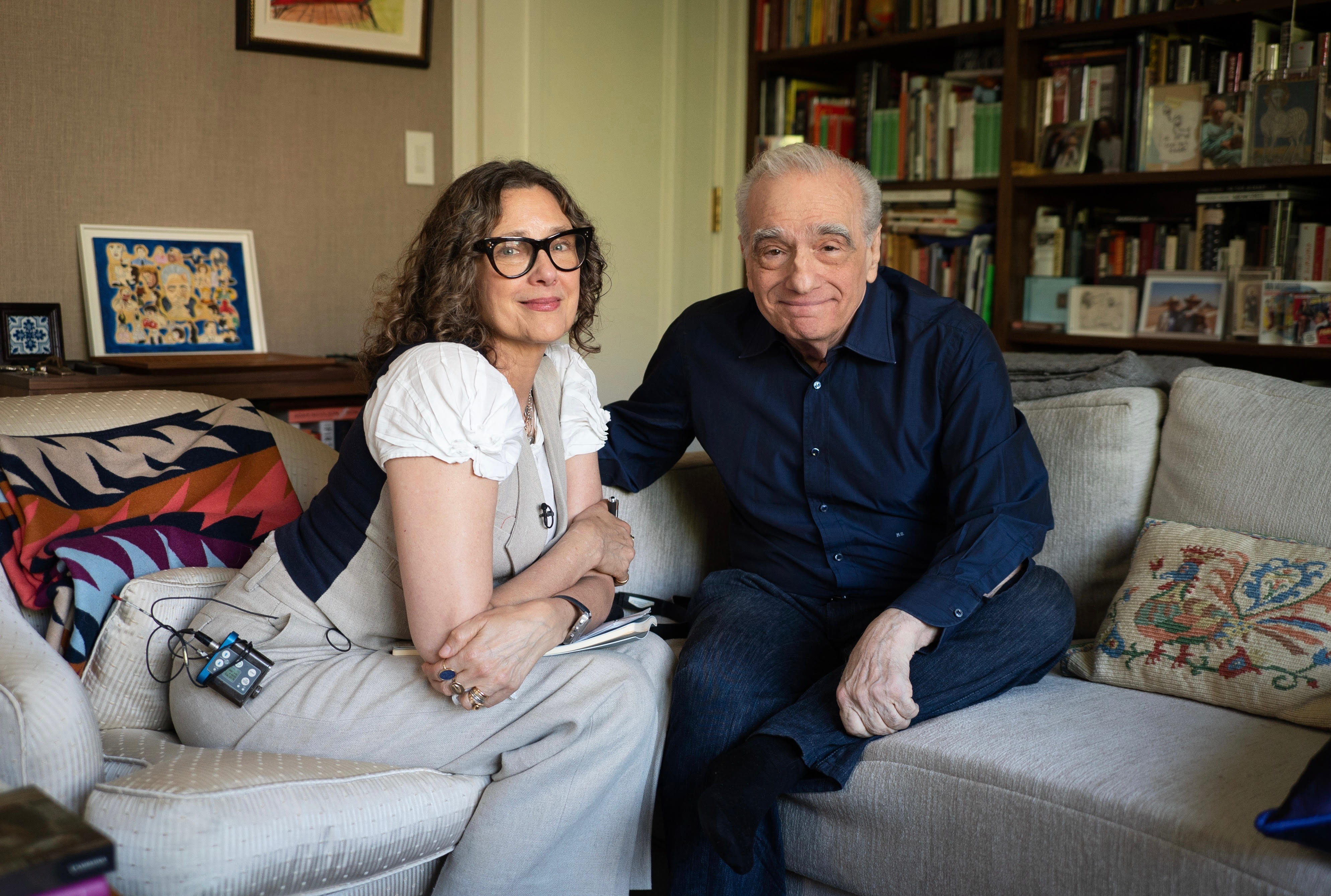 In a living room, a woman and a man wearing lavalier mics sit on a gray couch.