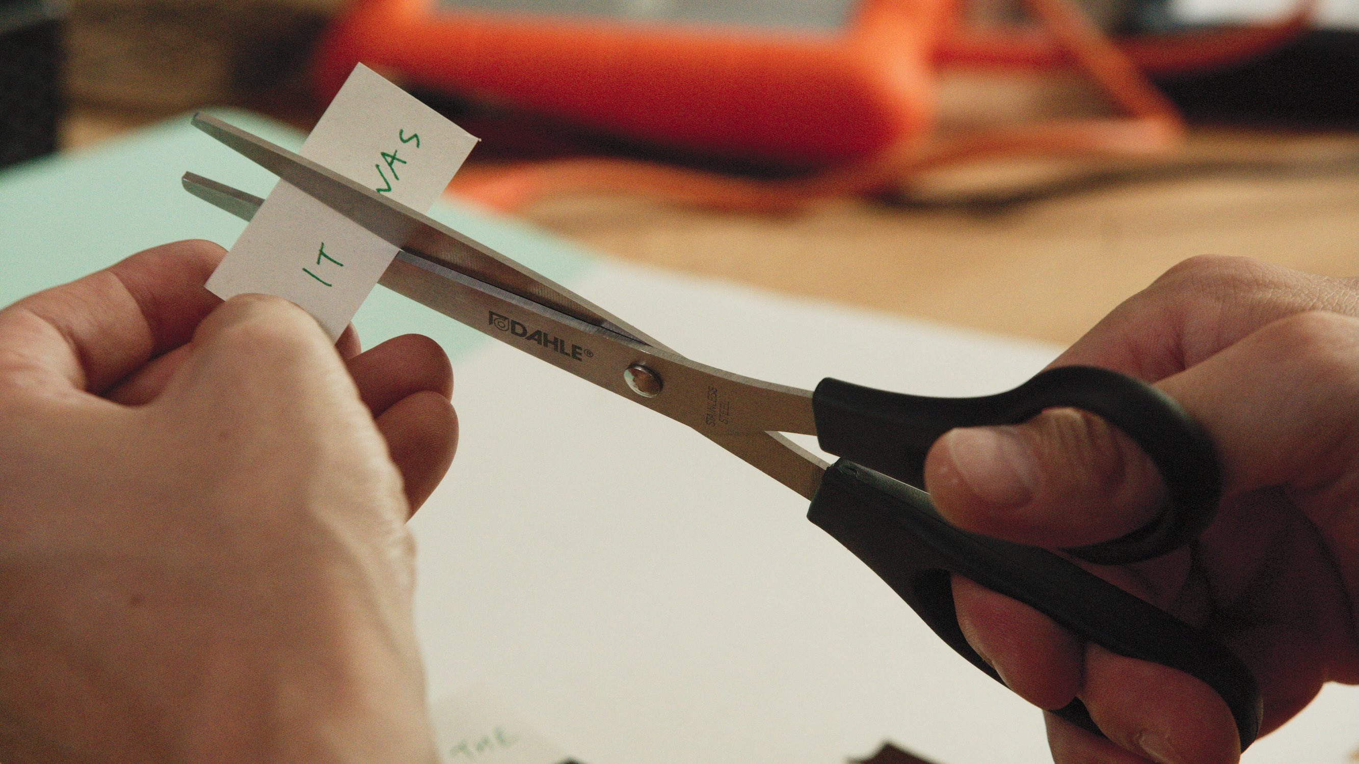 Closeup of a person cutting a piece of paper with "It was" written on it in half with a pair of black scissors.
