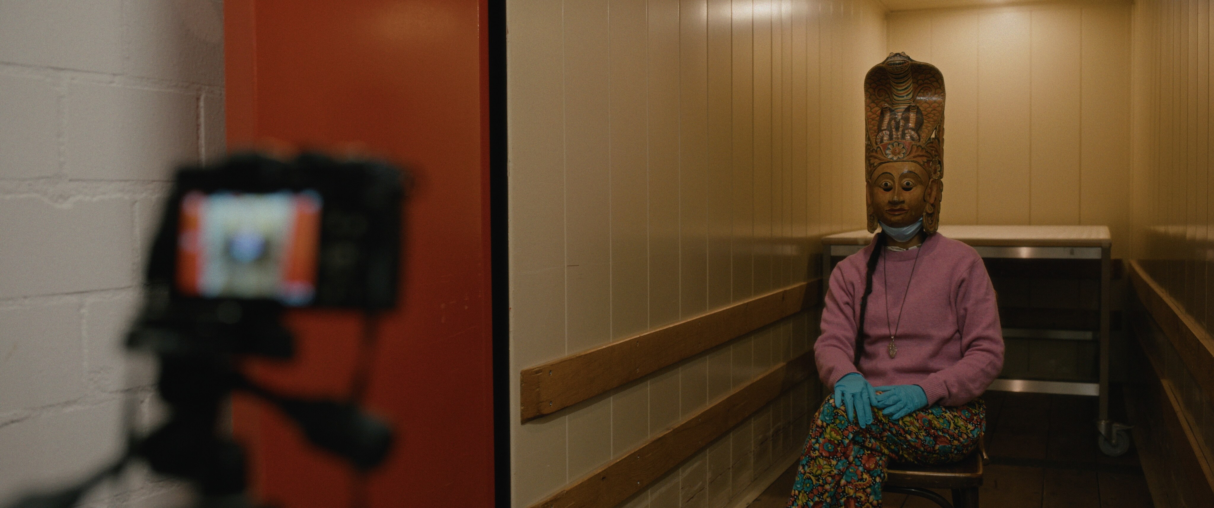 A camera on a tripod captures a portrait of a woman wearing a pink sweater, latex gloves, and a wooden indigenous mask in a nondescript room.