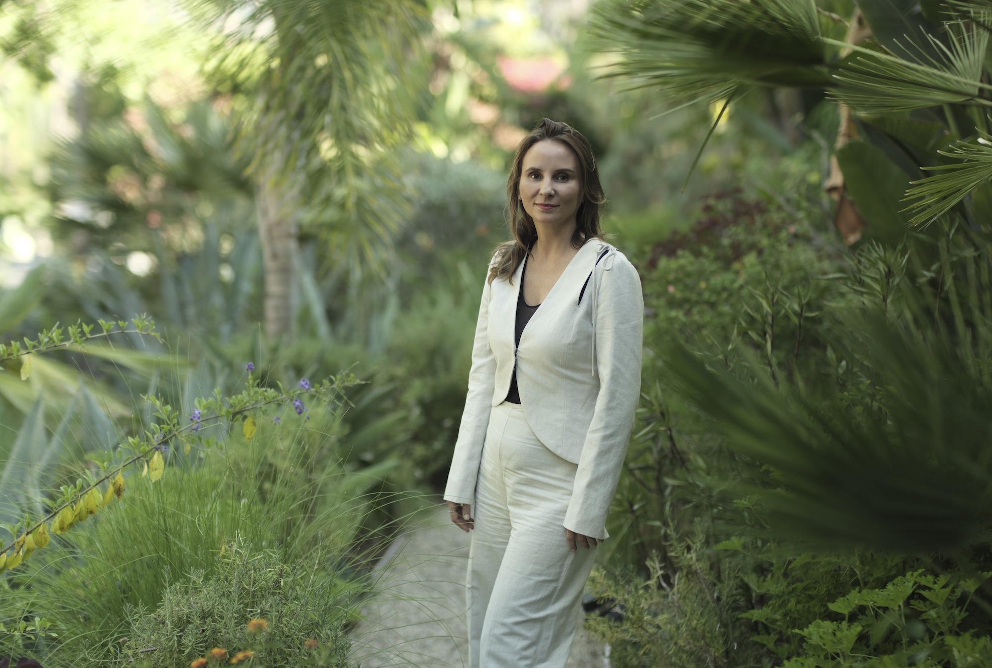 Woman with shoulder length brown hair in a white suit stands against a lush green garden backdrop.