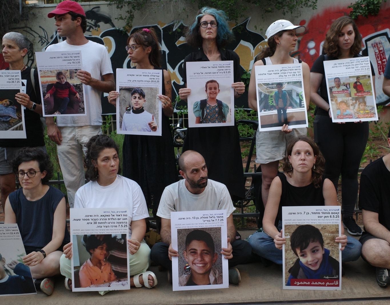 Two rows of people stand in silent vigil outside, holding large print-out posters of children's faces.