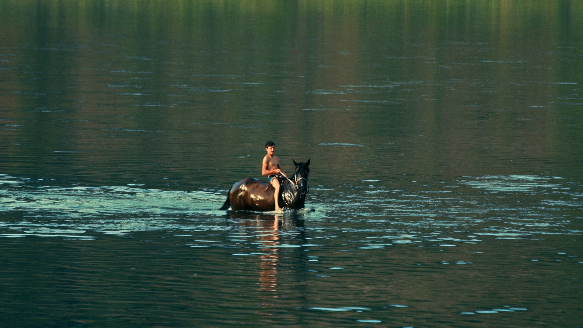 A drenched young man in nothing but a swimsuit rides a horse in the middle of a body of water
