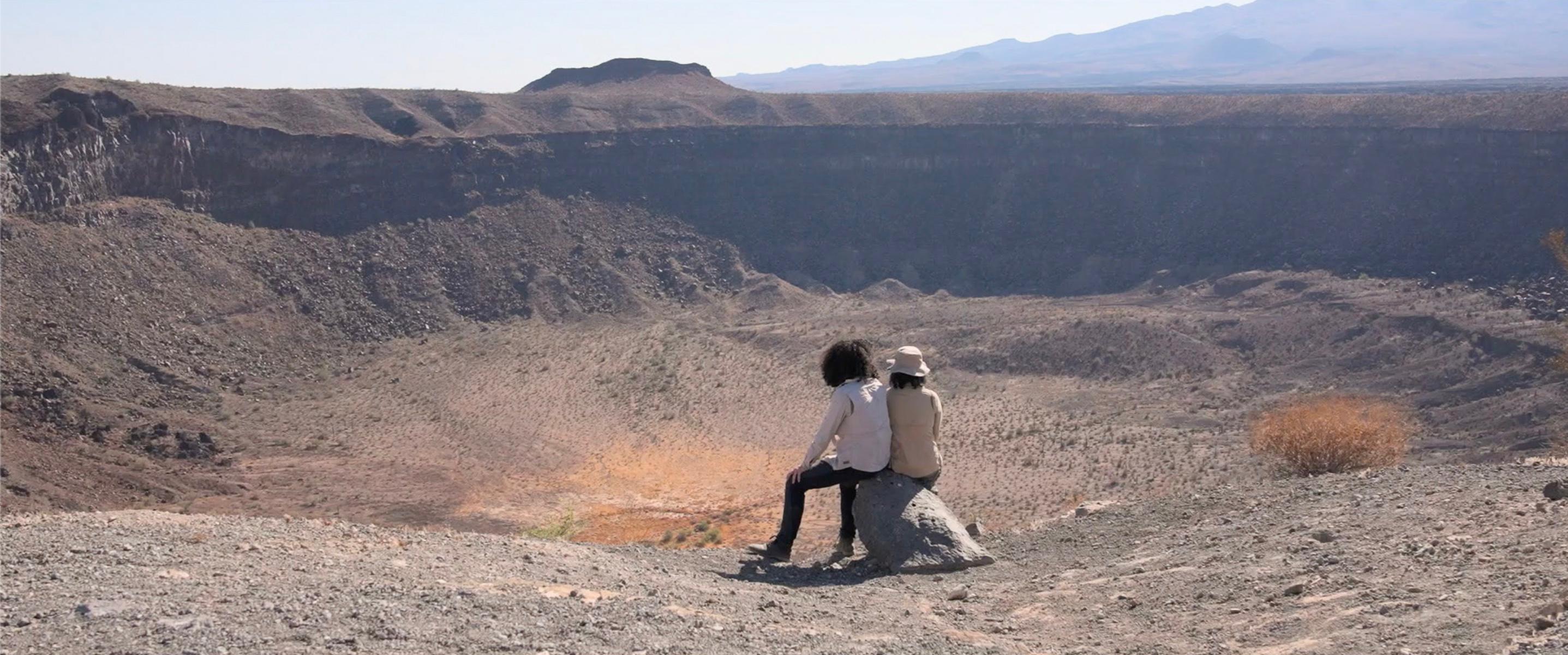 Two people sit on a rock against a vast desert landscape before them
