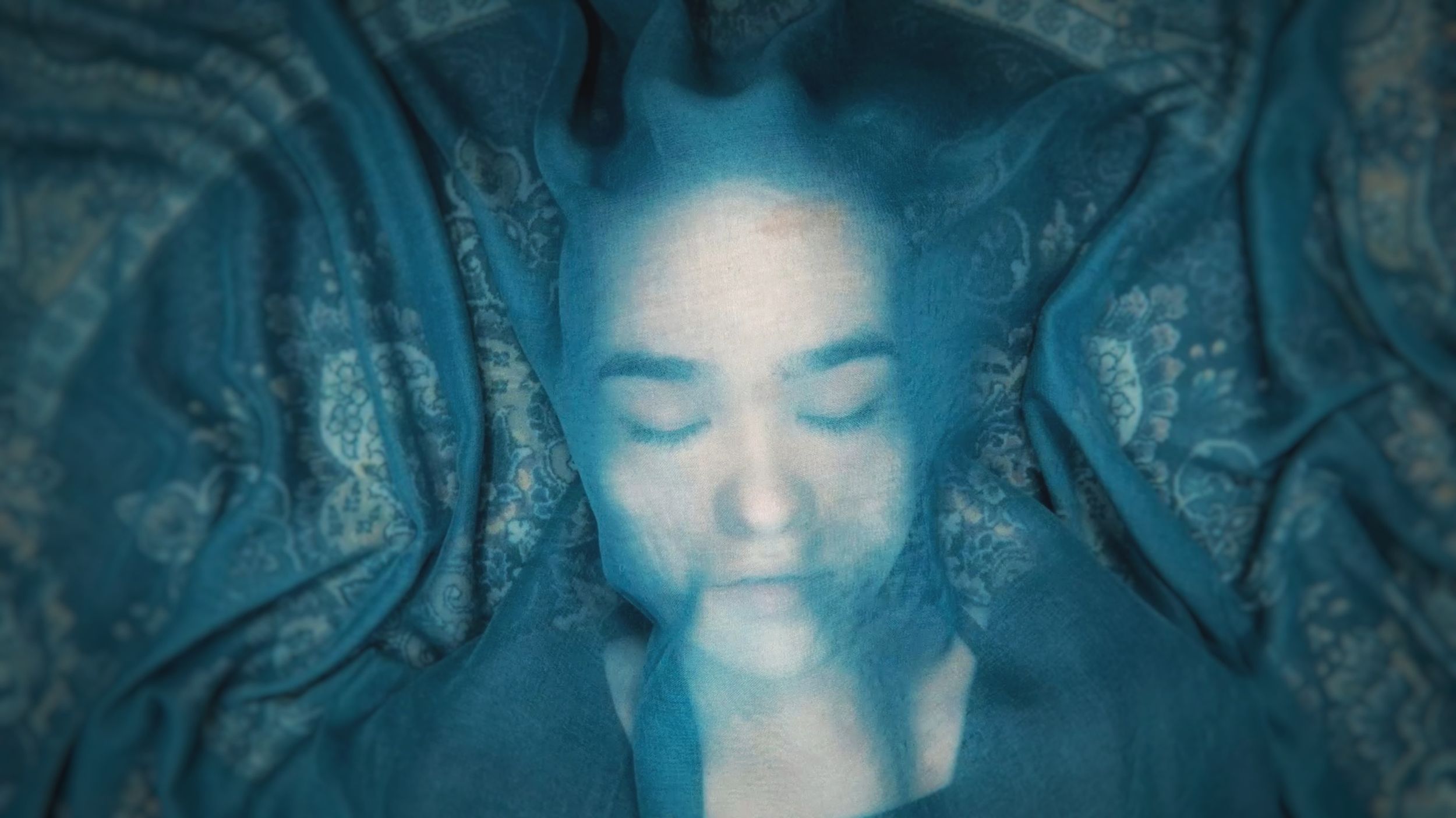 Under a blue cloth that is semi-transparent, a teenaged girl lays on the floor with her eyes closed.