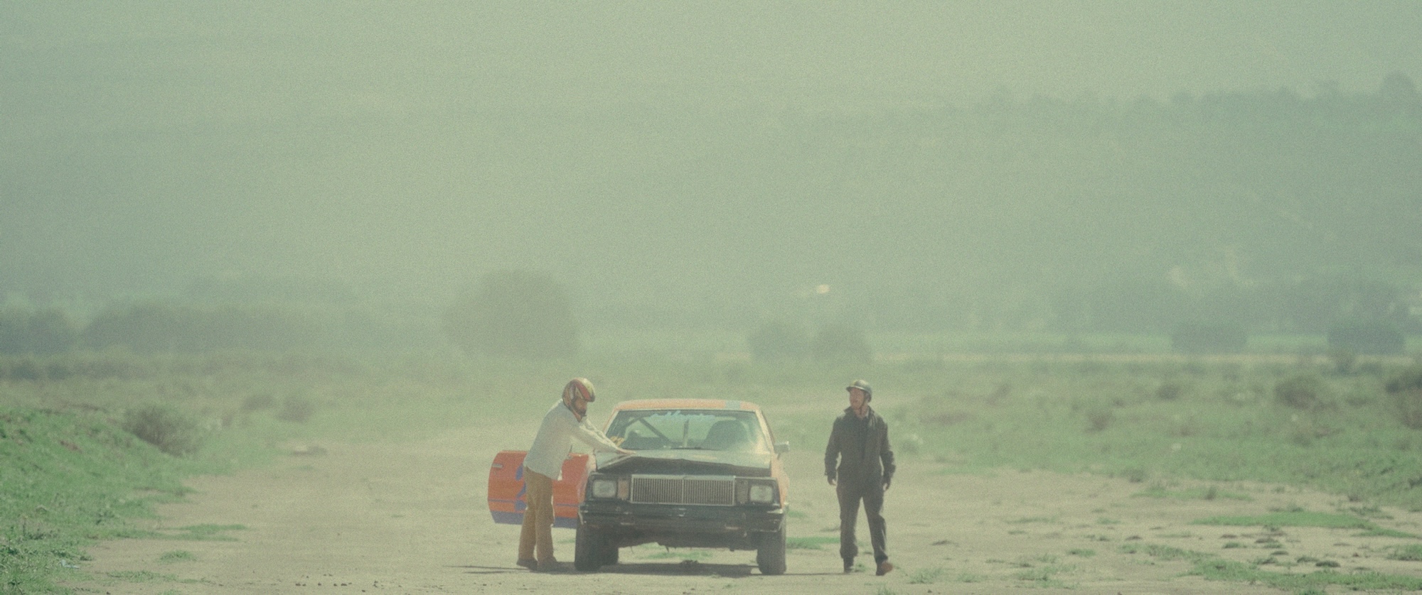 In a dusty road, two men with helmets on stand on either side of a broken down, battered car.