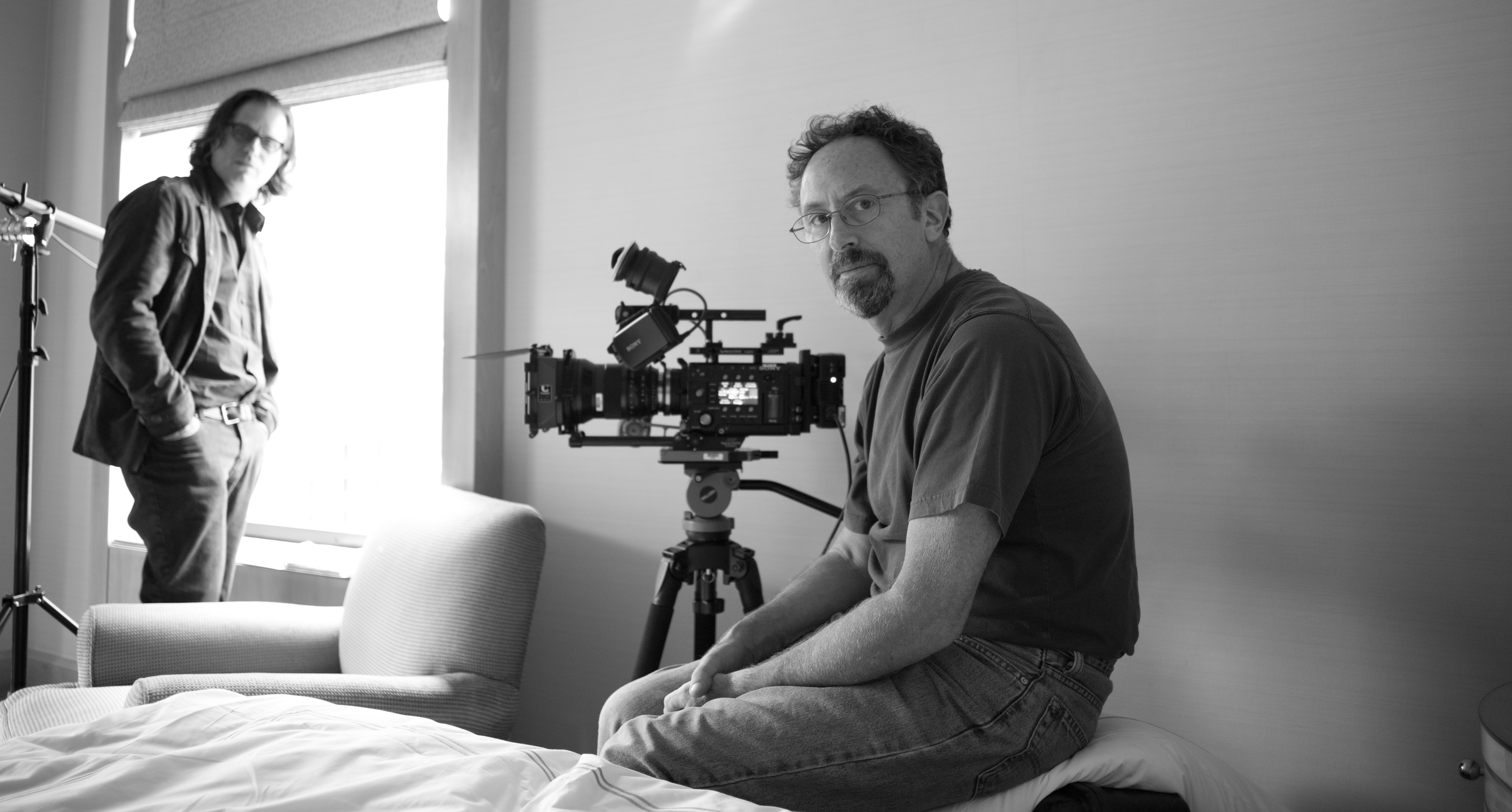 Black and white image of a white man with short hair, glasses and goatee, man sitting in a room next to a camera, as another stands by a window.