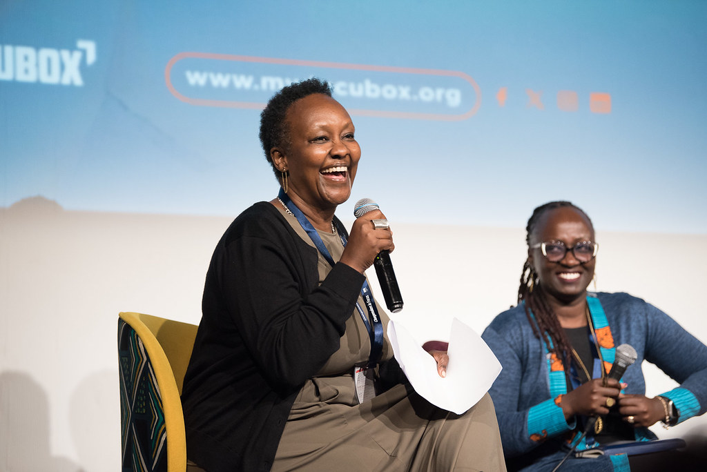 A Black woman with short-cropped hair sits at a stage next to another Black woman; both are holding mics and smiling widely