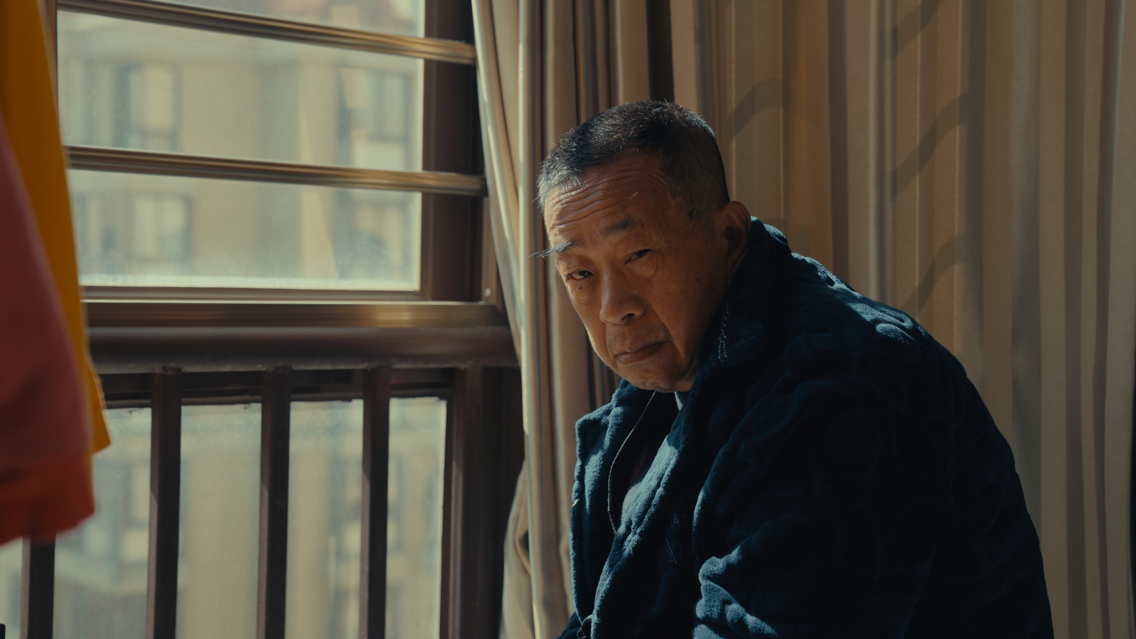 An aging Asian man with short-cropped hair in a bulky winter jacket glares at the camera while hunched over