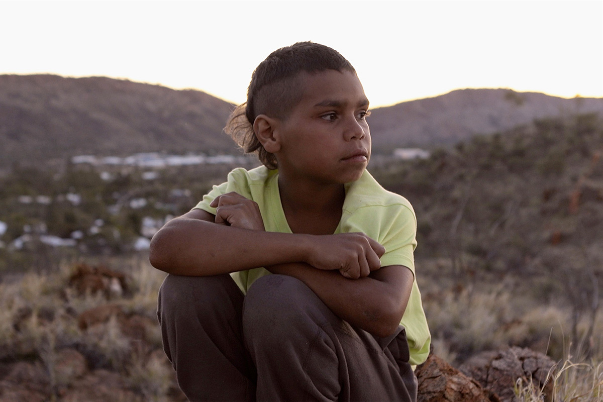 A young Dujuan Hoosan is sitting on a hill, looking pensively to his left. He is from the Arrernte Nation, group of Aboriginal Australian peoples from Central Australia.