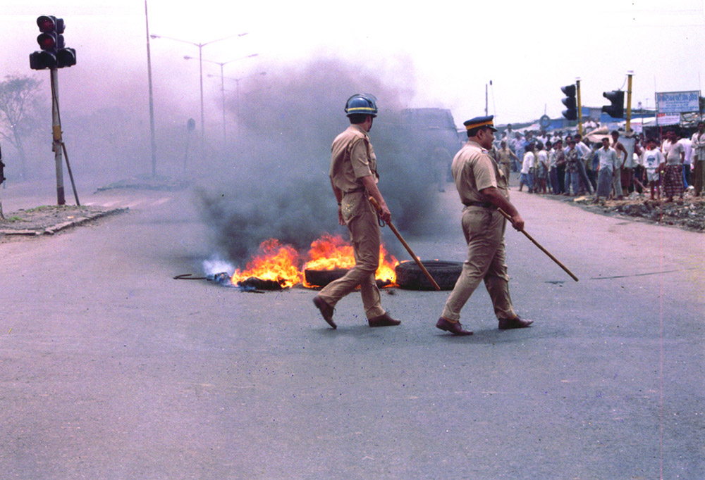 Tires burning in the middle of the road, two Indian police officers are walking by with batons in hand and crowd watching on the side of the road. Courtesy of OVID.tv