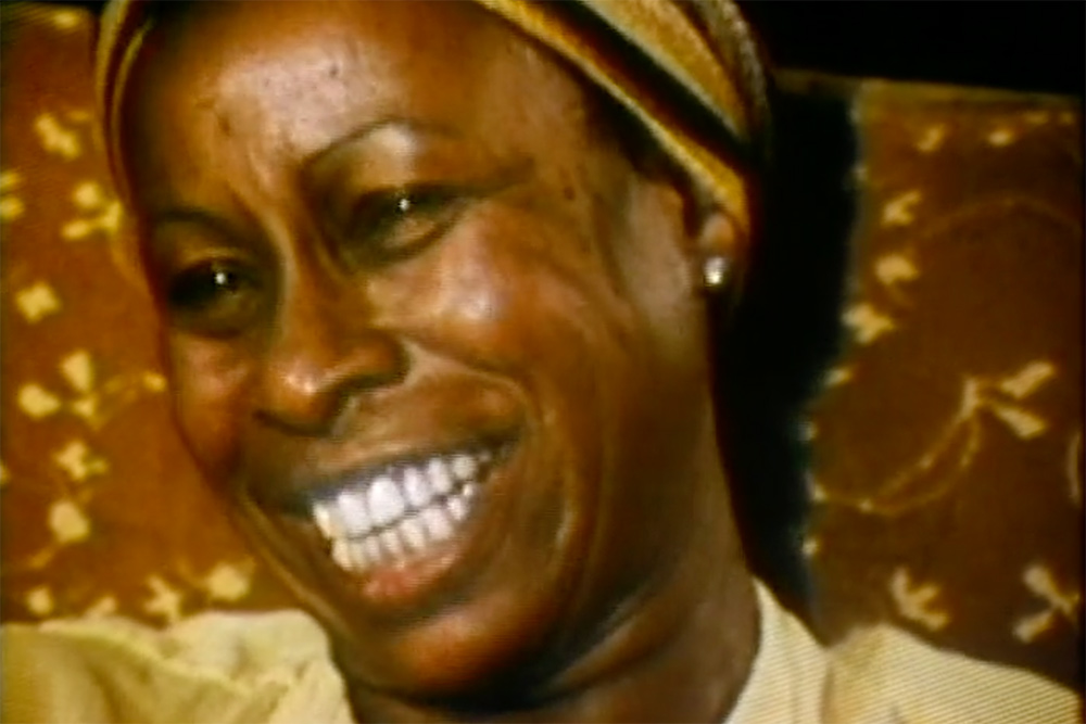 An image of Betty Carter, a Black female jazz singer. She is smiling, wearing a golden yellow blouse and a headband. From Michelle Parkerson’s “. . . But Then, She’s Betty Carter”. Courtesy of The Criterion Collection.