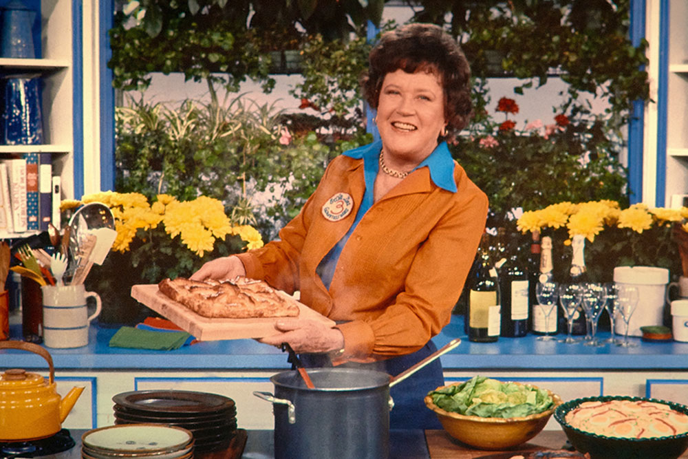 Julia Child is a white chef with short brown wavy hair. Here she is standing in her kitchen holding a tray of pastry. She is wearing a blue shirt underneath another brown one, and is smiling. Image from Julie Cohen and Betsy West’s ‘Julia.’ Courtesy of TIFF.