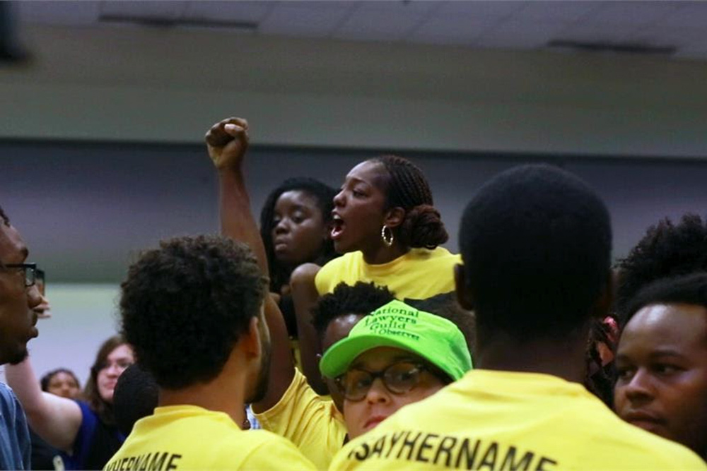 Caption: Janaé Bonsu is a young Black woman leading an activist gathering. She and her comrades are in yellow shirts that say “Say Her Name”. Image for Ashley O’Shay’s “Unapologetic”. Courtesy of David Magdael & Associates.