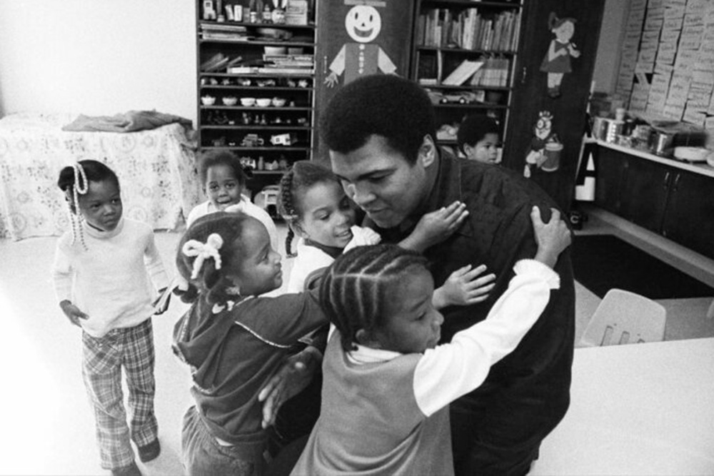 Muhammad Ali, a Black boxer, on a visit to his old grammar school in Louisville, KY, in 1977. He is surrounded by young Black children who are hugging him. From ‘Muhammad Ali,’ a four-part documentary from Ken Burns, Sarah Burns and David McMahon. Photo by Michael Gaffney. Courtesy of PBS.