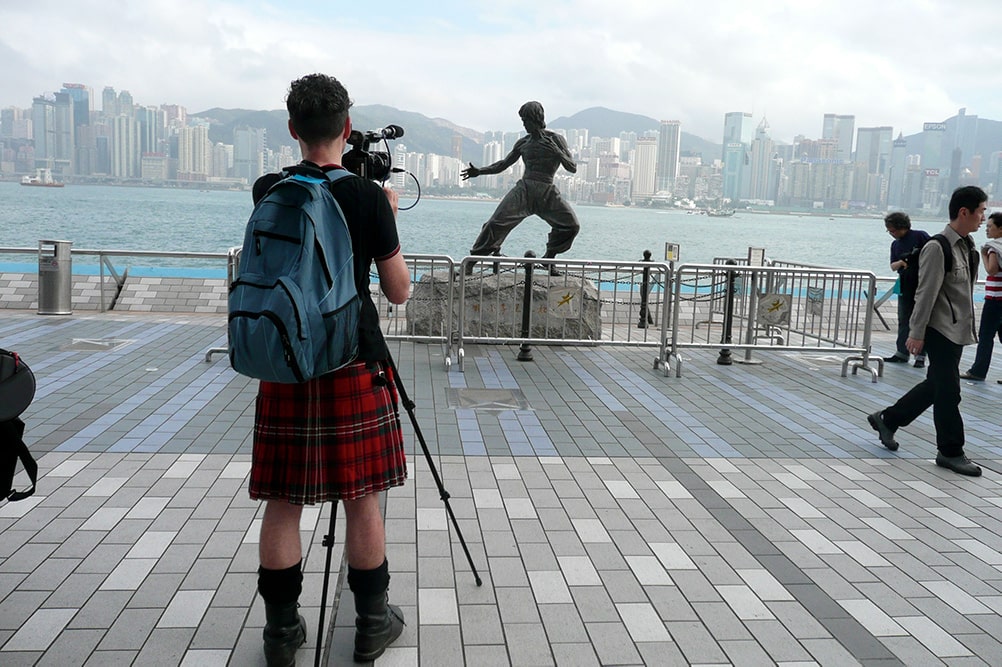 A cameraperson wearing a kilt filming a Bruce Lee statue in Hong Kong. From Mark Cousins’ ‘The Story of Film: An Odyssey’. Courtesy of Music Box Films.