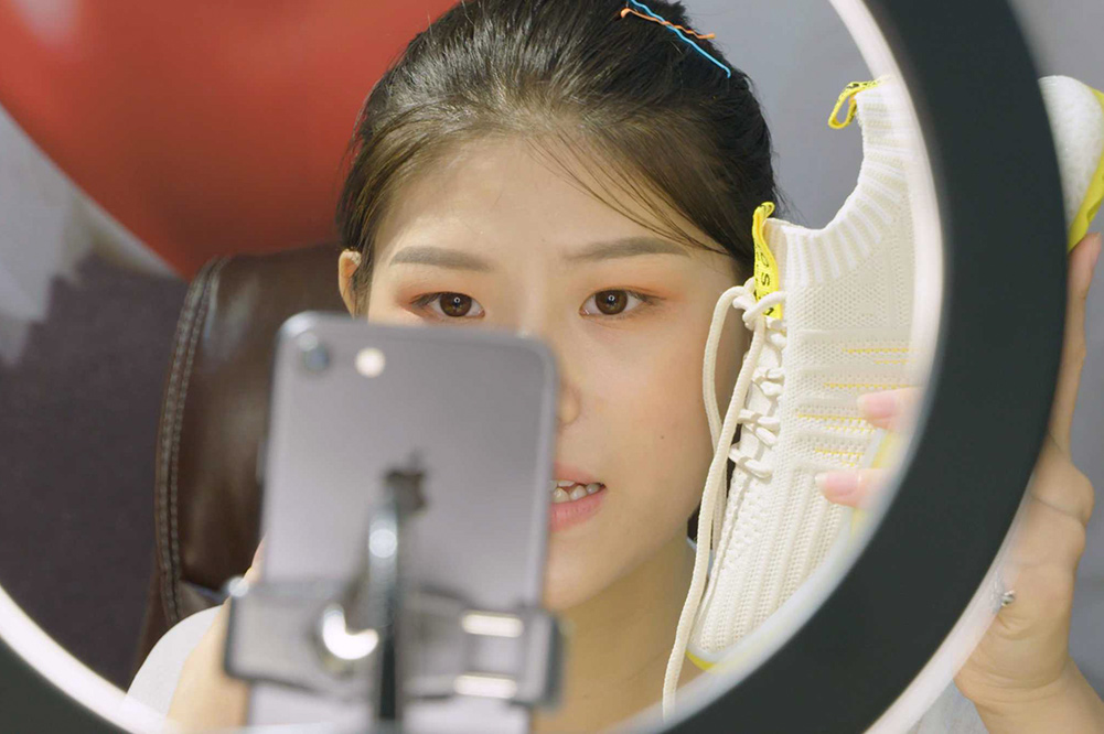A young Chinese woman recording herself on an iPhone, holding a sneaker. There is a ring light in front of her. Image from Jessica Kingdon’s “Ascension.” Courtesy of Tribeca  Festival