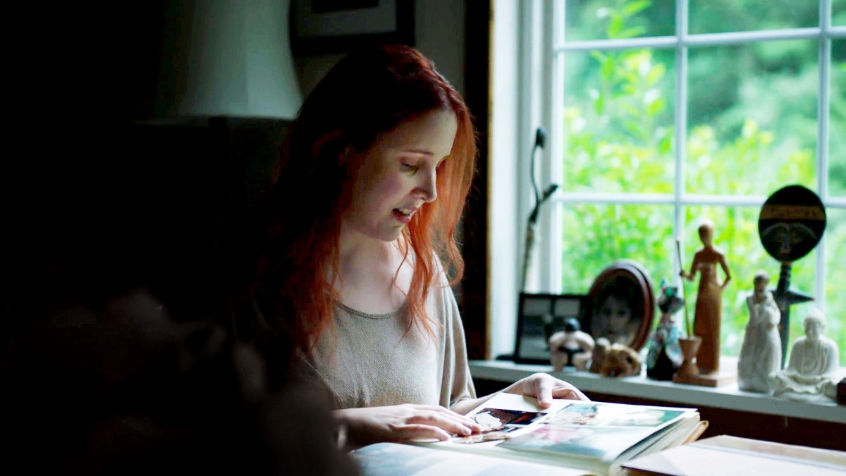 Dylan is a white woman, with red hair, in her early 30s; she is sitting at a desk next to a window, and she is looking at a photo album.