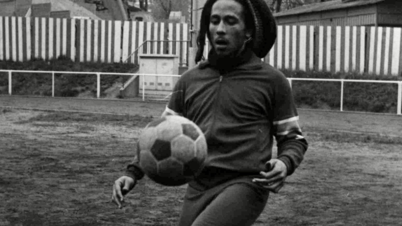 Reggae legend and football fan Bob Marley, in dreadlocks and beard and wearing a dark jersey, dribbles a football. From ‘Legacy:Rhythm of the Game.’
