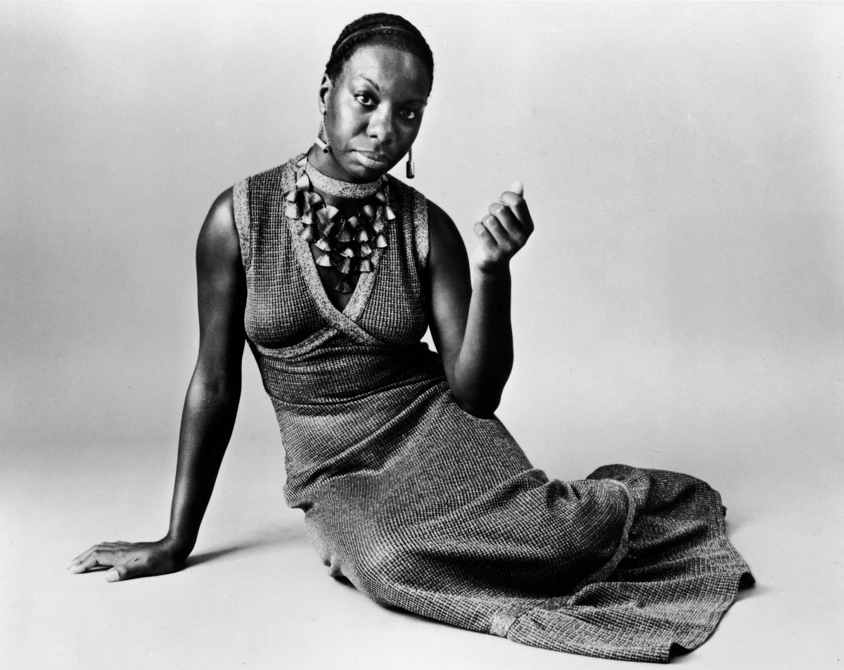 Nina Simone is sitting on the floor with her right arm propping her up. She is wearing a maxi knit dress, large decorative necklace.