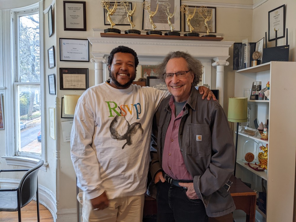 Amir George (left) , the new artistic director of Kartemquin Films, with his predecessor, Founder/Artistic Director Gordon Quinn (right). Amir is a Black man with a beard, wearing a white sweatshirt, and Gordon is a white man with gray hair and glasses, wearing a grey coat  over a purple button-down shirt. . Photo courtesy of Anthony Kauffman