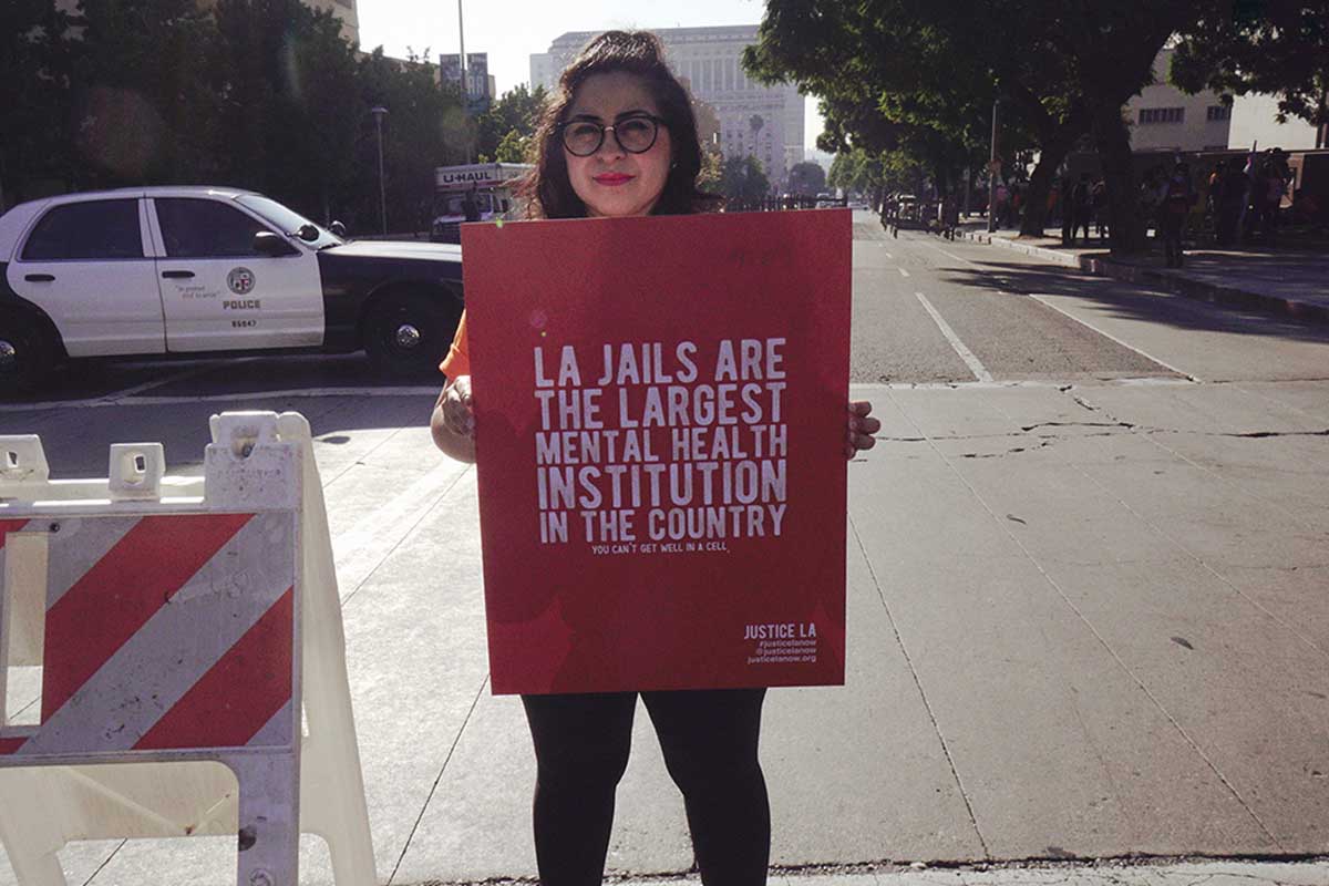​A woman wearing a hat and glasses, with shoulder-length hair, stands on a street and holds a sign that reads "LA Jails are the largest mental health institutions in the country." From Kenneth Paul Rosenberg’s 'Bedlam.' Courtesy of 'Independent Lens.'