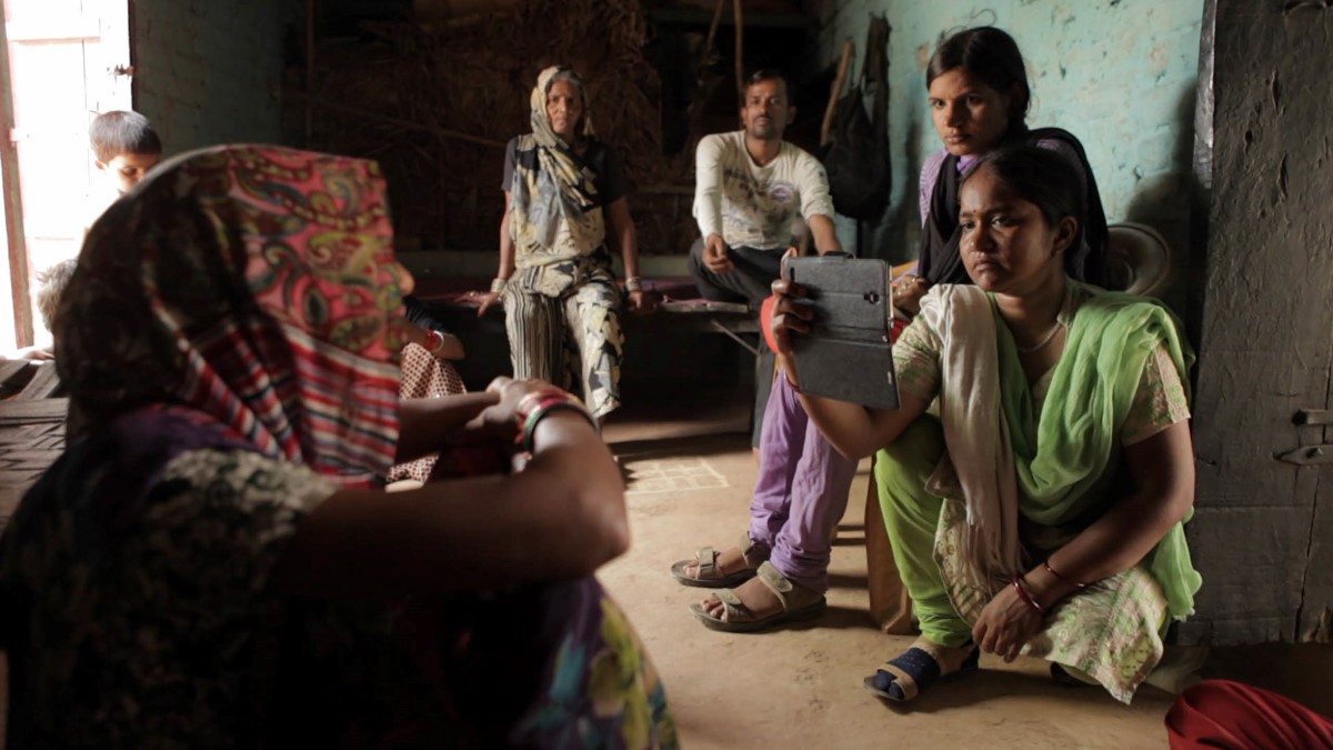 At right, A reporter from an India-based digital platform is kneeling while interviewing an Indian woman who is seated at left, in her living space with her family. The reporter is filming with a smartphone in her right hand , nd is wearing an emerald green sari