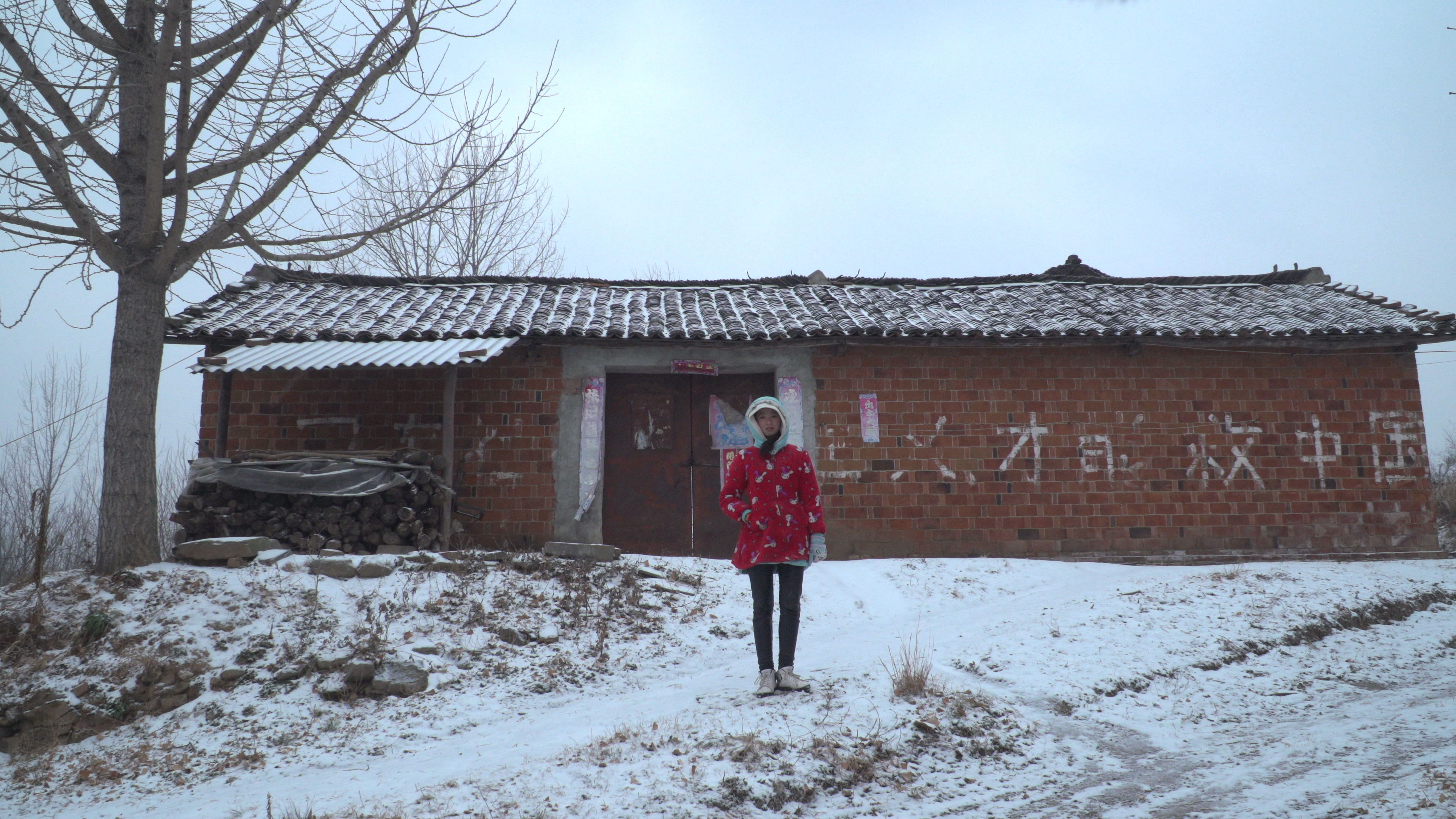 A young girl in a red coat stands in front of a brick building in the middle of winter.