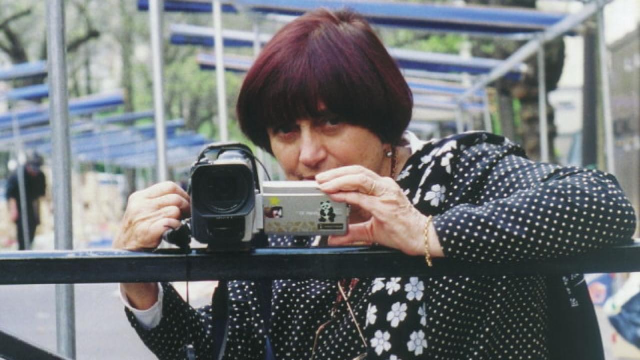 Agnes Varda holds a videocamera with stickers on it up against a railing.