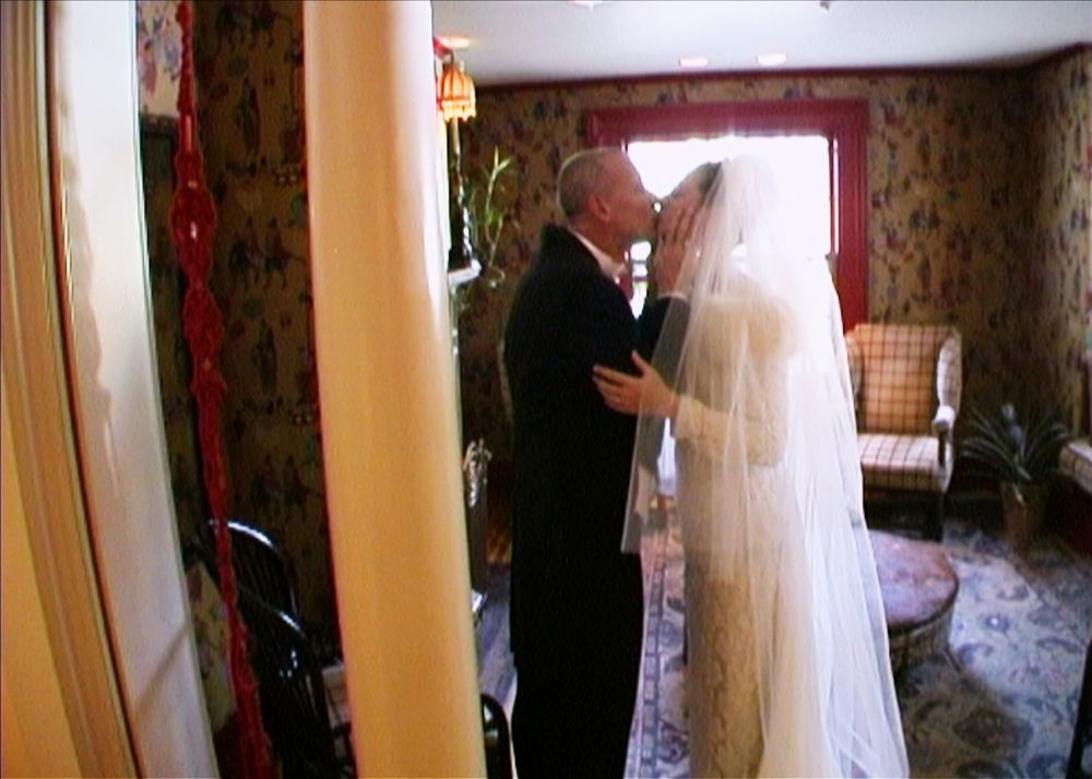 A man and woman, dressed in a tuxedo and a wedding gown and veil, kiss in a living room