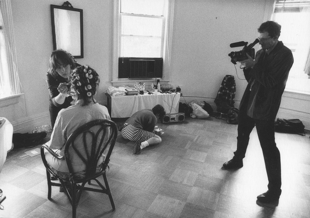 Black and white photo of a woman with her hair in curlers getting her makeup done, a woman writing on a piece of paper on the ground, and man videotaping