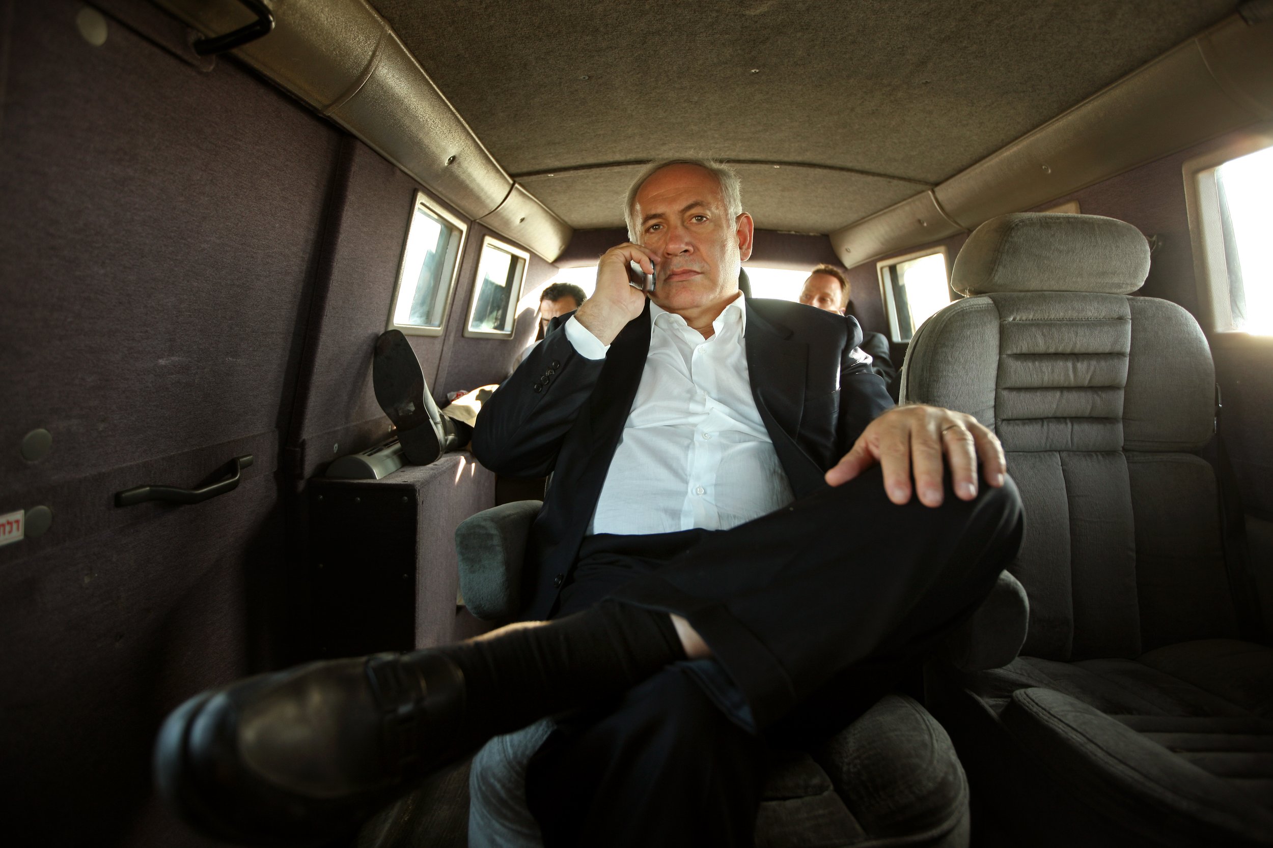 Inside a car, a gray-haired man in a black suit holds a cell phone up to his ear.