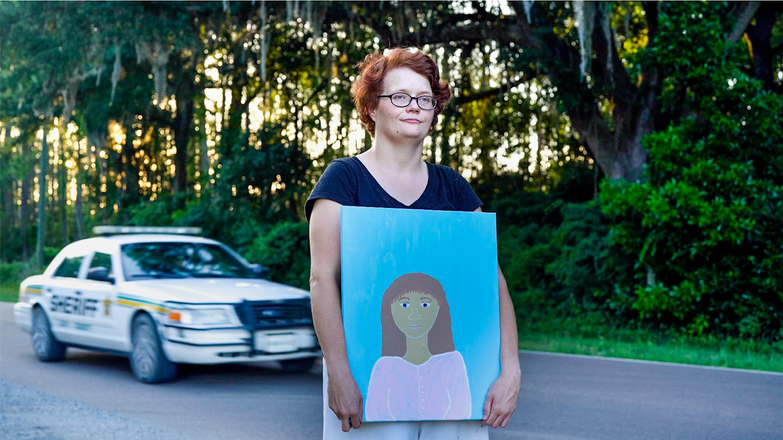 White woman with short red hair and glasses holds in front of her body a painted self-portrait of herself as a young girl while a police car drives behind her on a rural highway. Sun is glimmering in through trees in the background. In the painting she is wearing a pink shirt and is in front of a light blue background.