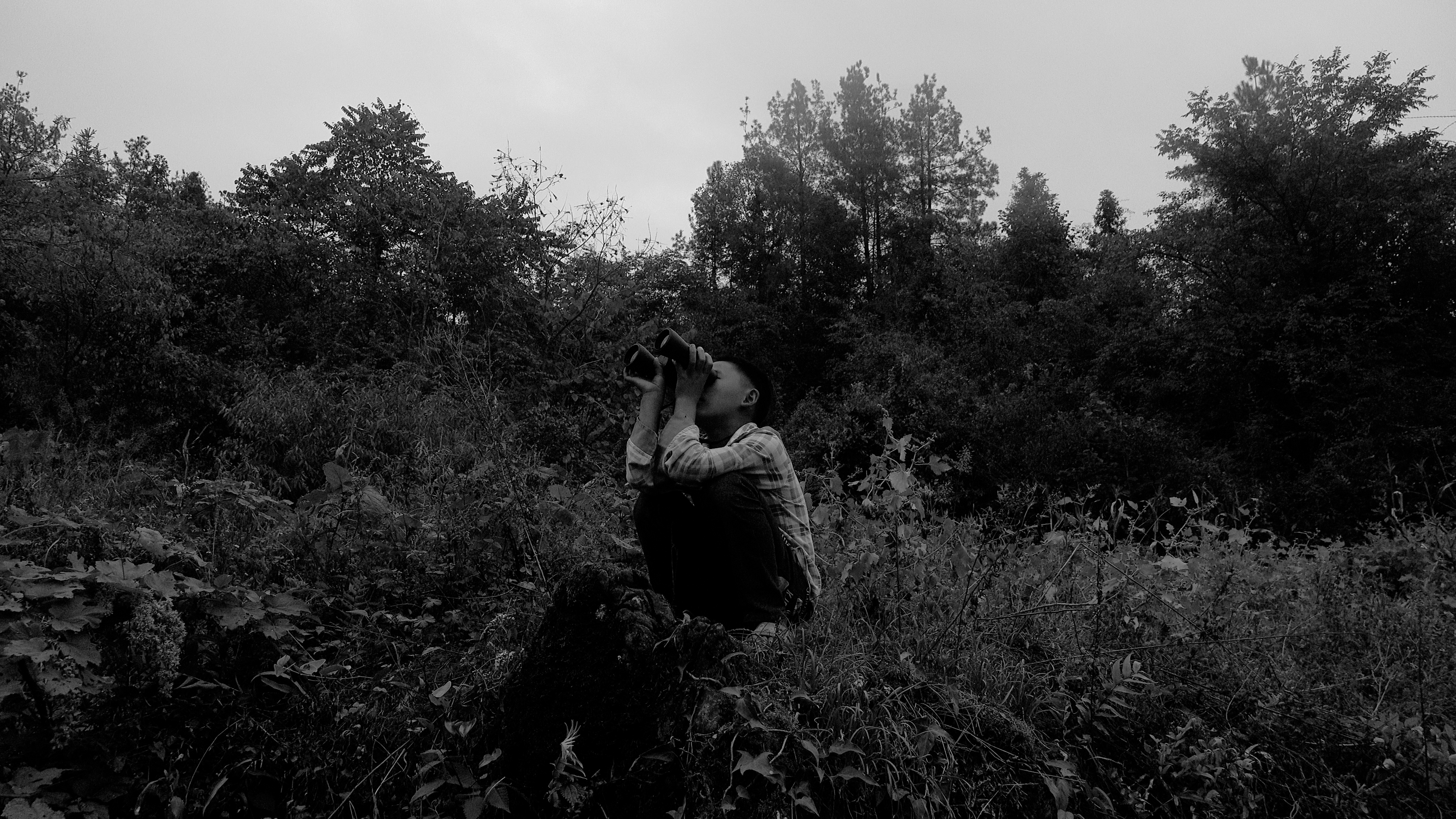 Black and white image of a person squatting in a field, looking through binoculars.