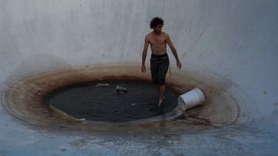 A young man, shirtless with curly hair, stands in the dirty water at the bottom of an empty pool