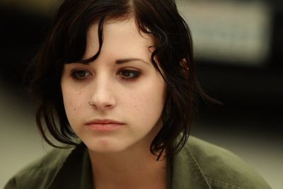 A young woman with dark hair framing her face sits and stares down into the distance, frowning