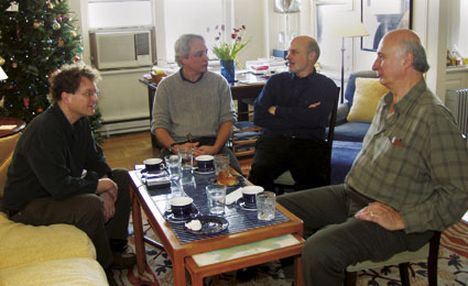 Four white men, middle-to-older aged, sit around a coffee table in a living in conversation 