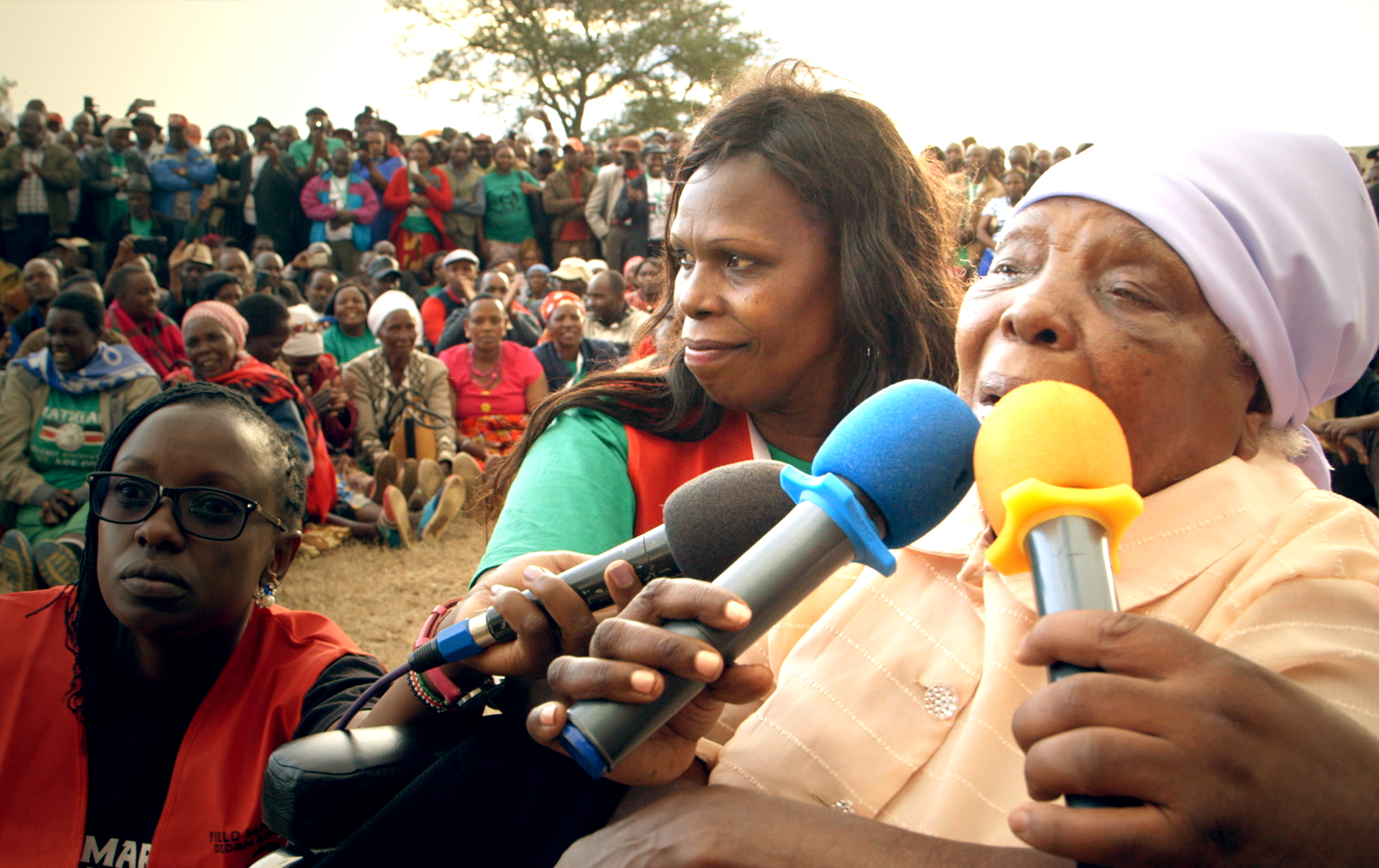 A film still from Our Land, Our Freedom that shows an elderly Black woman surrounded by several microphones, her face a picture of determination as she is mid-sentence. She wears a purple hat, and beside her are two women in red vests, also holding microphones. Behind them, is a large group of Black individuals, some standing and others sitting, focusing intently on the elderly woman as she speaks.