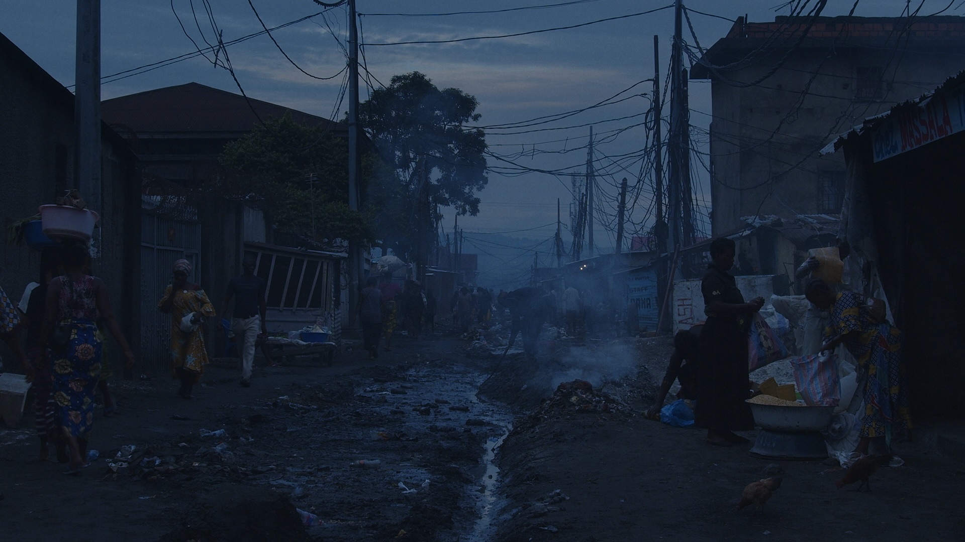A film still from Rising Up At Night that shows a disheveled rural African neighborhood in the dawn light. There are several little shops and people wearing traditional attire conducting errands, crossing over the wet dirt and trash littered roads.